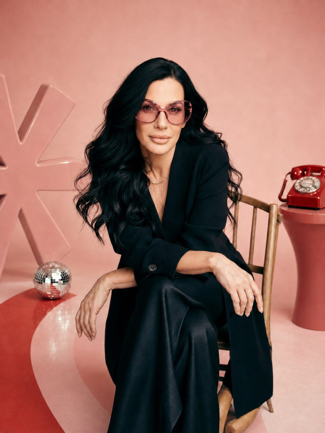 Brea Ballard seated in black tailoring and pink sunglasses beside the House of Icon asterisk, disco ball and red telephone